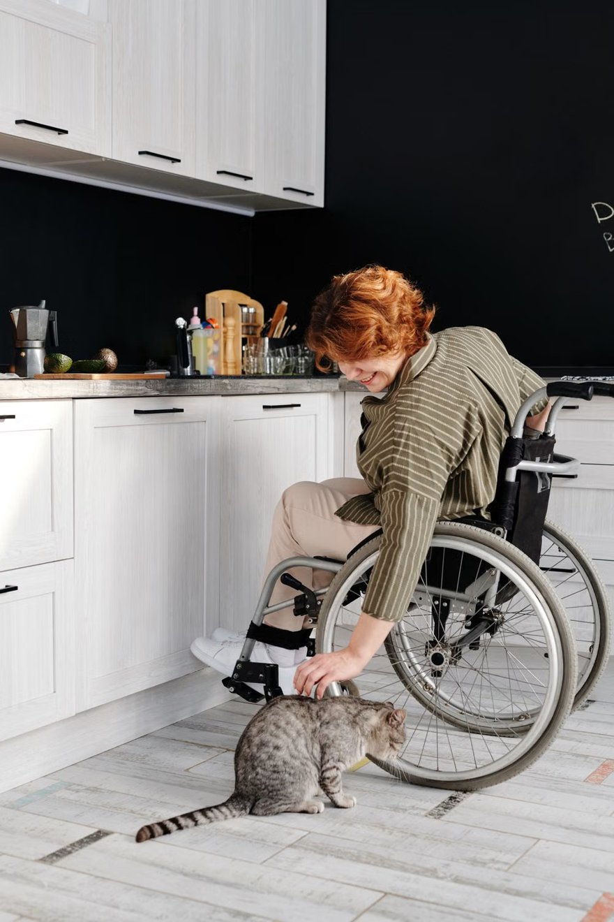 A woman in a wheelchair in her kitchen, reaching down to pat a cat